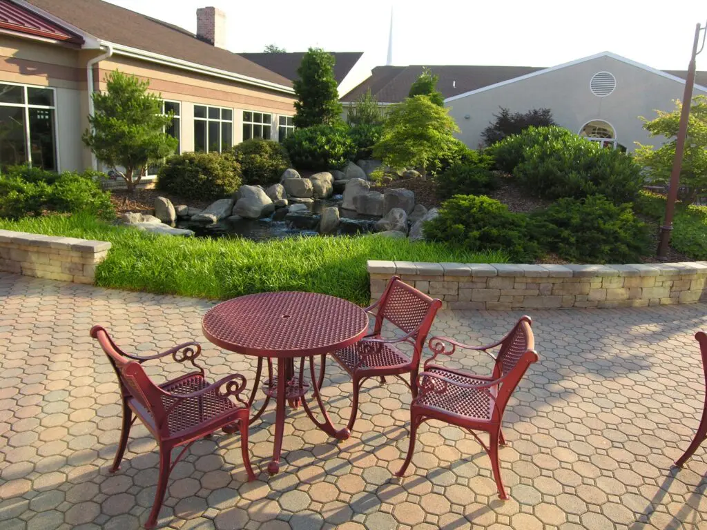 A Patio With A Round Red Metal Table And Three Matching Chairs Is In The Foreground. The Patio Is Paved With Hexagonal Bricks. In The Background, There Is A Small Landscaped Area With A Rock-Filled Water Feature, Various Plants, And A Building With Large Windows.