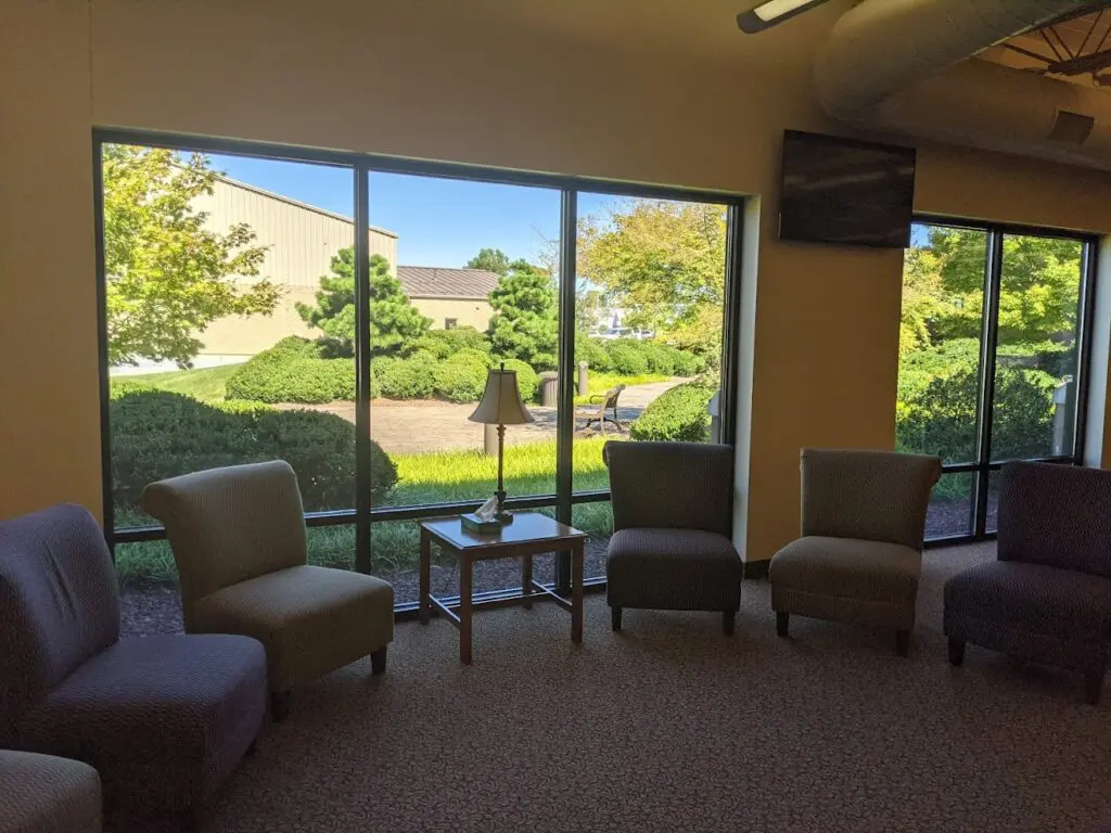 A Waiting Area With Several Cushioned Chairs Arranged In A Semicircle Around A Small Table With A Lamp. Large Windows Offer A View Of A Sunny Outdoor Scene With Trees And Bushes. A Television Is Mounted Near The Ceiling In The Corner.