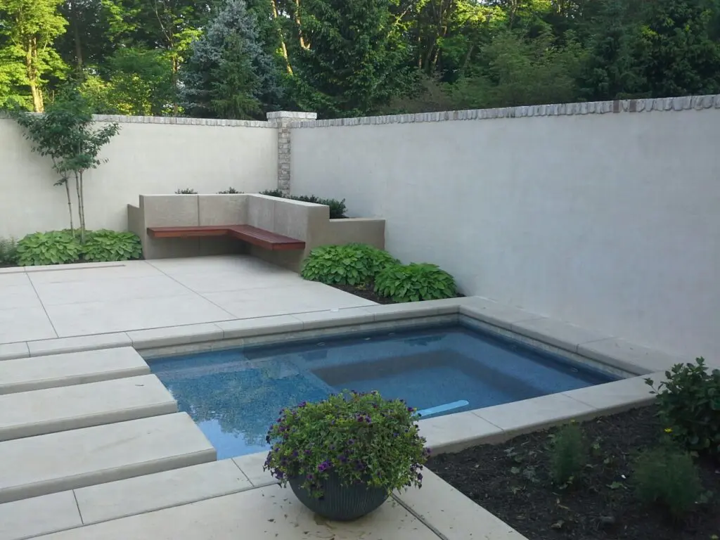 A Small, Minimalistic Backyard Pool Area With Modern Concrete Pavers And A Built-In Wooden Bench. The Pool Is Surrounded By A Simple White Wall And Various Green Plants, While A Pot With Purple Flowers Decorates The Corner. Trees Are Visible In The Background.