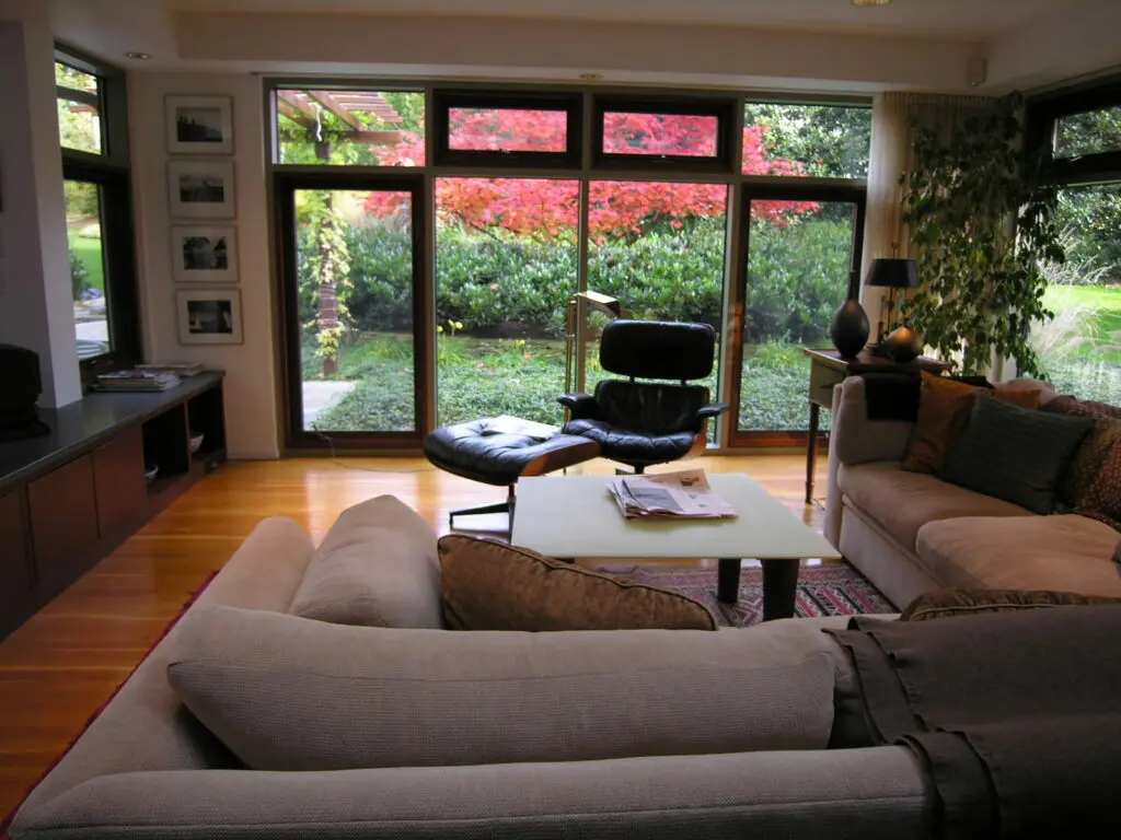 &Quot;Inside Out&Quot; Gardens 16 A Cozy Living Room Features Modern Furniture, Including A Sectional Sofa, Coffee Table With Newspapers, And A Black Lounge Chair With An Ottoman. Large Windows Showcase A Vibrant Garden With Colorful Foliage, Bringing Natural Light And Greenery Into The Space.
