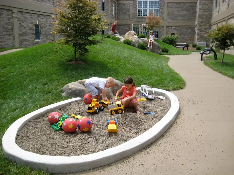 Two Children Are Playing With Toy Trucks And Balls In A Sandbox Area Surrounded By Grass And Small Trees. A Paved Path Winds Through The Park-Like Setting, Leading To A Multi-Story Brick Building. Other Children Are Playing And Walking Around In The Background.