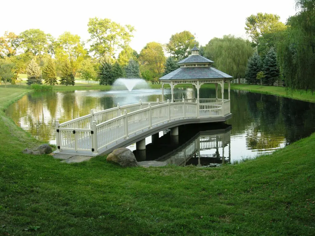 Garden Structures 8 A Serene Park Scene Featuring A White Gazebo At The Edge Of A Pond With A Wooden Bridge Leading To It. A Fountain Sprays Water In The Middle Of The Pond, Surrounded By Lush Green Grass And Trees In The Background.