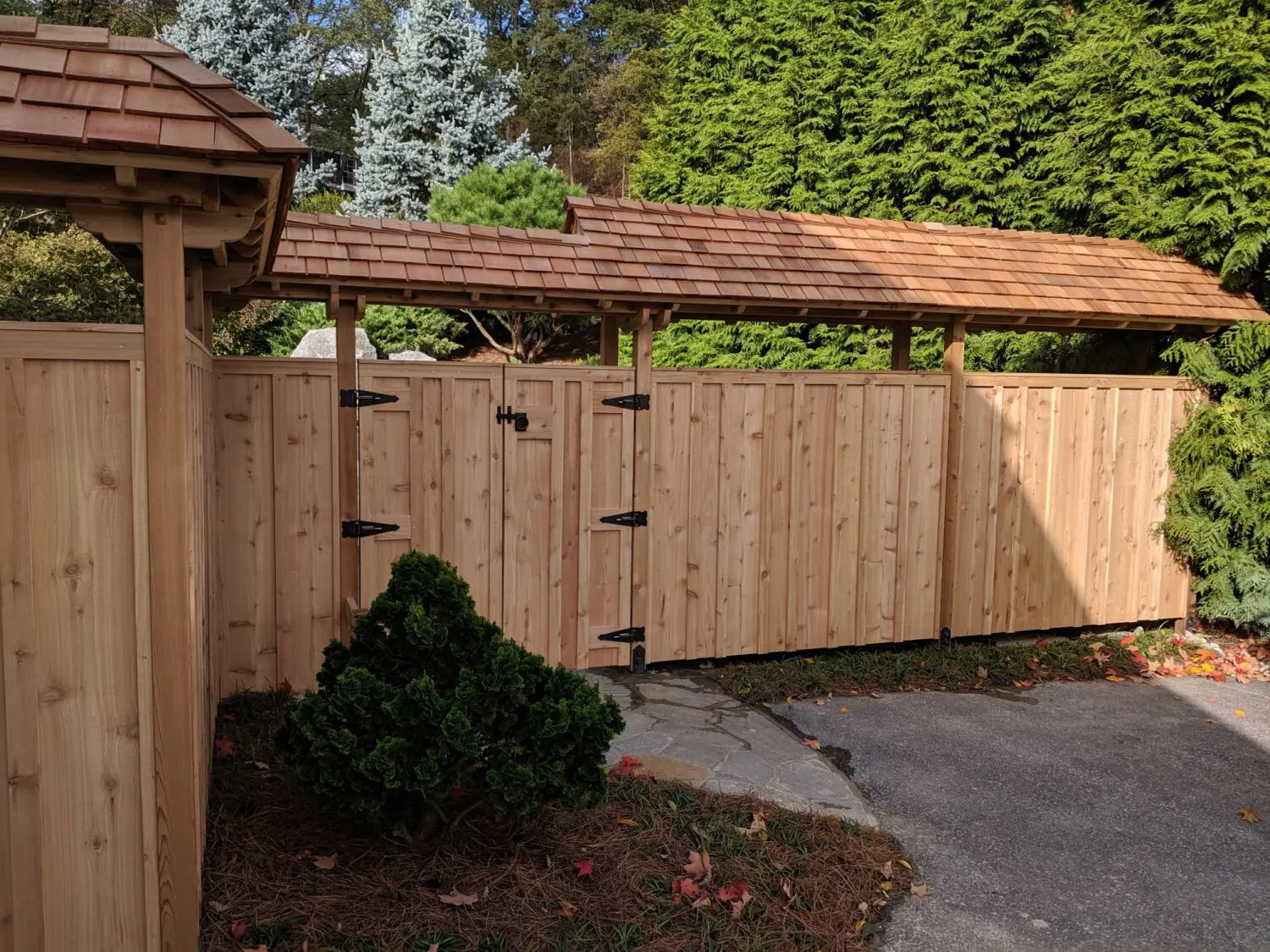 A Wooden Fence And Gate With A Natural Finish Stand Alongside A Landscaped Garden. The Gate Features Black Metal Hinges, And There Is A Small Wooden Gazebo To The Left. Evergreen Trees And Shrubs, Including A Small Conifer, Provide A Lush Green Backdrop.