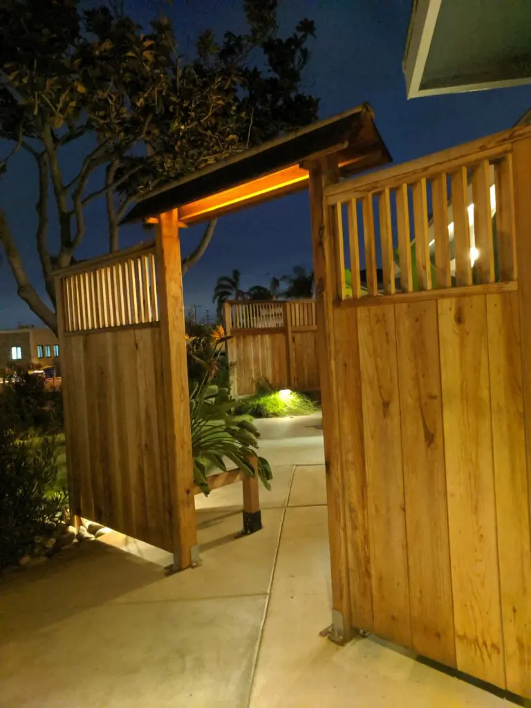 Garden Structures 11 A Wooden Gate Warmly Lit From Above Stands Open, Framed By A Wooden Fence. The Paved Pathway Illuminated By Lights Leads Into A Landscaped Garden Area With Trees And Foliage Visible In The Background Under A Dusky Sky.