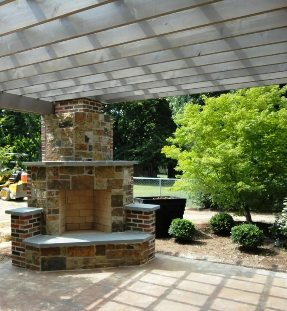 Garden Structures 14 A Rustic Outdoor Stone Fireplace Underneath A Wooden Pergola. The Fireplace Features A Brick And Stone Facade With Built-In Seating. The Area Is Set Within A Well-Manicured Garden With Lush Green Plants, A Tree, And A Fence In The Background.