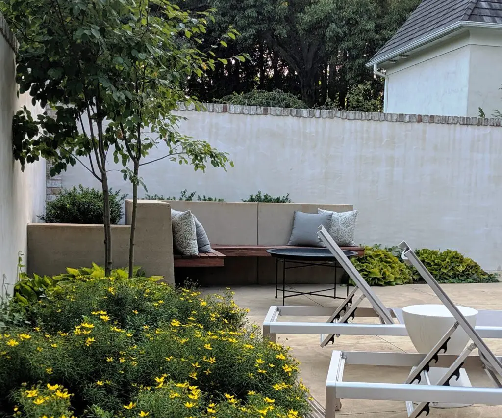 Garden Structures 15 A Cozy Outdoor Patio Area Featuring A Wooden Bench With Grey Cushions, A Small Round Table, And White Lounge Chairs. A Lush Garden Bed With Yellow Flowers Is In The Foreground, And A Tall Wooden Fence With Green Foliage Is In The Background.