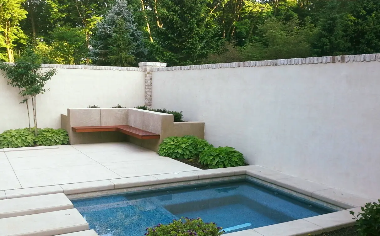 Low-Maintenance Gardens 16 A Minimalist Backyard Features A Small, Rectangular Pool With A Stone Border, Surrounded By A Concrete Patio. A Wooden Bench Is Nestled In A Corner With Beige Walls And Green Plants At The Base. Tall Trees And Bushes Provide A Lush Backdrop.
