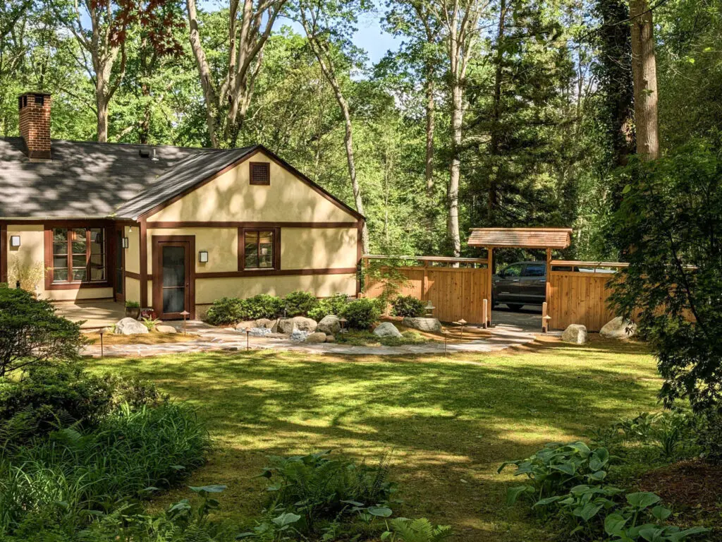 A Small Cottage-Style Home With Tan Walls, Brown Trim, And A Stone Walkway, Surrounded By A Lush Green Lawn And Dense Forest. A Wooden Gate And Fence Stand To The Right, With A Vehicle Partially Visible Behind The Gate    