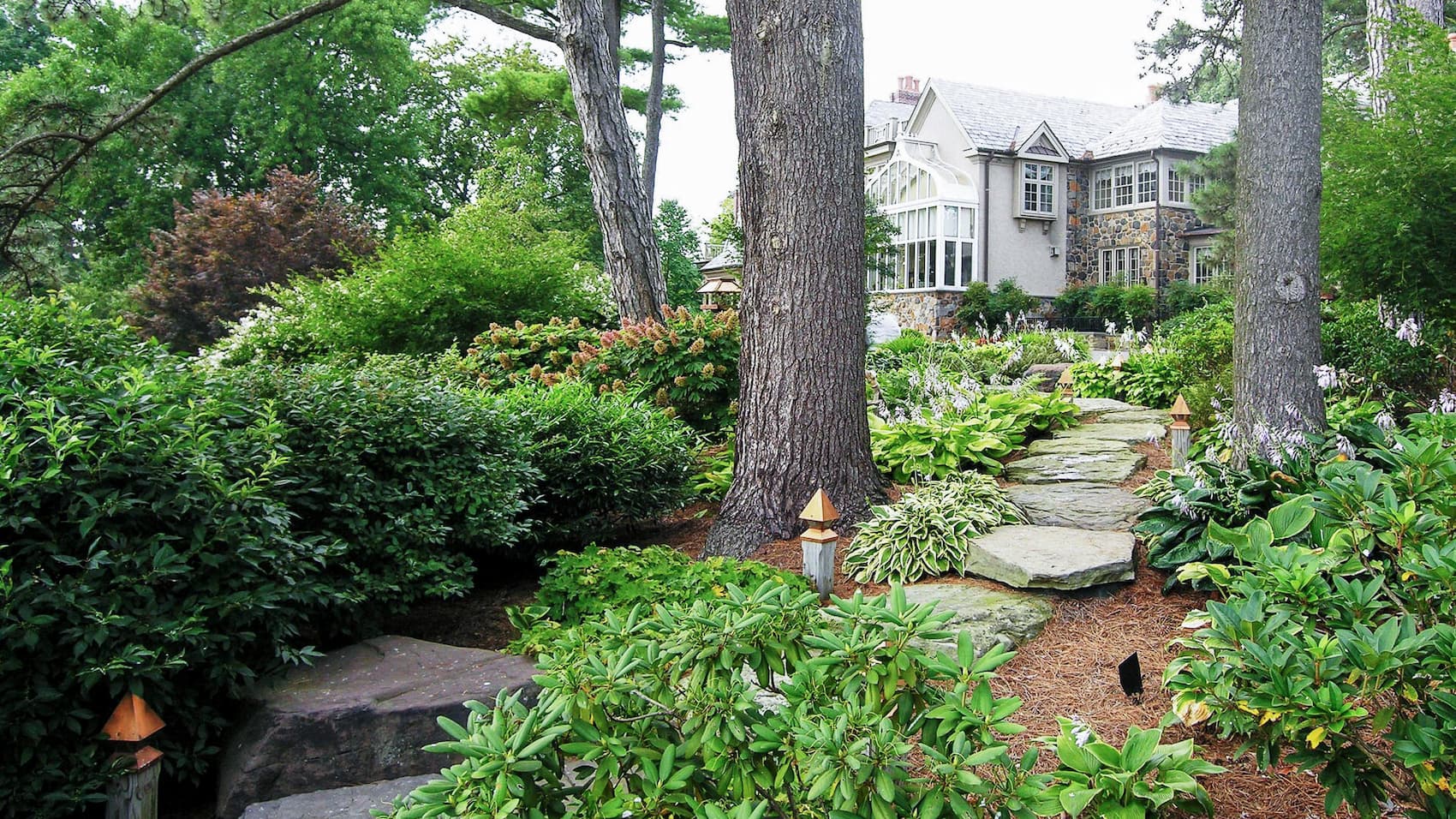 A Natural Stone Walkway Surrounded By Bushes, Tall Trees, And Walkway Lights