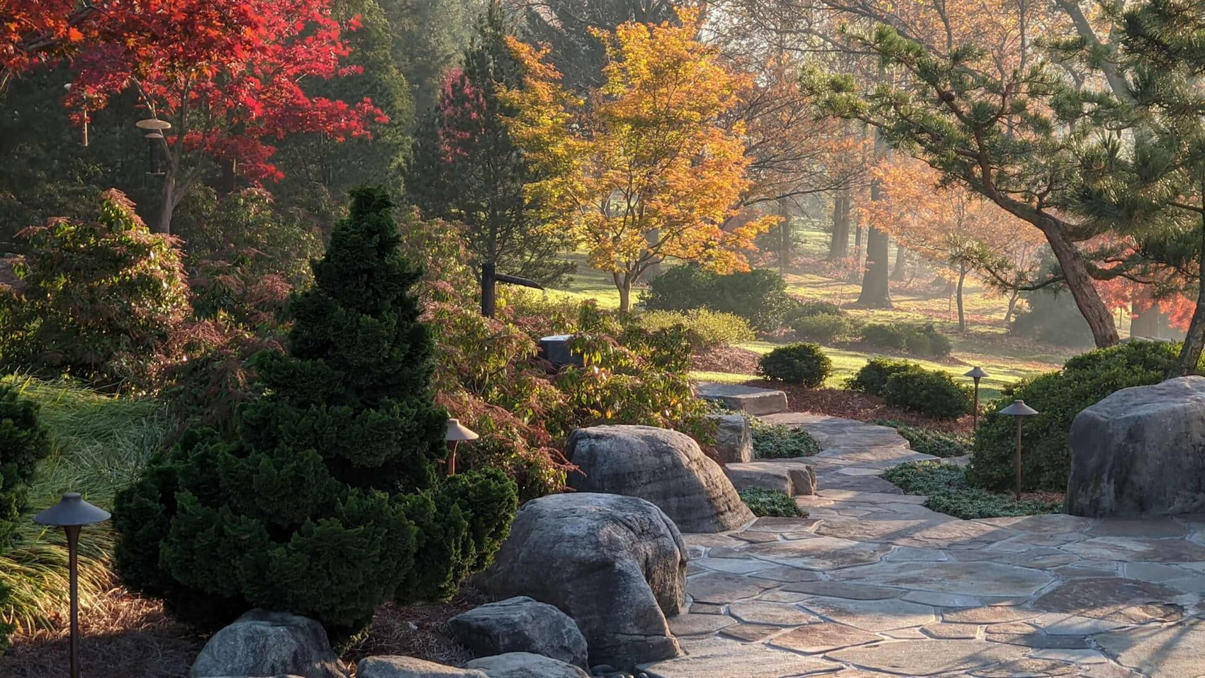 A Variety Of Trees And Well Maintained Bushes Surrounding A Stone Path That Leads Into A Lawn Manicured By Hanselman Landscape And Gardens